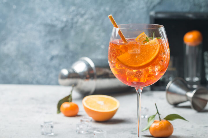 Ein großes Weinglas, gefüllt mit einem orangefarbenen WASGAU-Cocktail, Eiswürfeln und Orangenscheiben, garniert mit einem gestreiften Strohhalm. Auf dem Tisch liegen eine halbierte Orange, kleine Mandarinen mit Blättern, Eiswürfel und Barutensilien im Hintergrund.