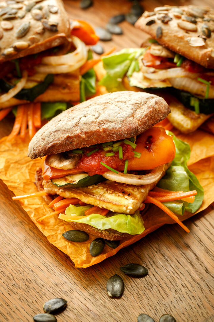 Drei gestapelte WASGAU Veggie-Sandwiches auf rustikalem Brot mit Salat, gegrilltem Gemüse, Karotten, Zwiebeln, Tomaten und grüner Beilage werden auf Pergamentpapier auf einer Holzunterlage mit gestreuten Kürbiskernen angerichtet.