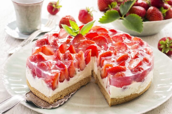 Ein köstlicher WASGAU Erdbeerkäsekuchen mit frischen Erdbeerscheiben und klarer Glasur liegt auf einem Teller, von dem eine Scheibe entfernt ist. Eine Schale mit Erdbeeren und eine Tasse stehen im Hintergrund.