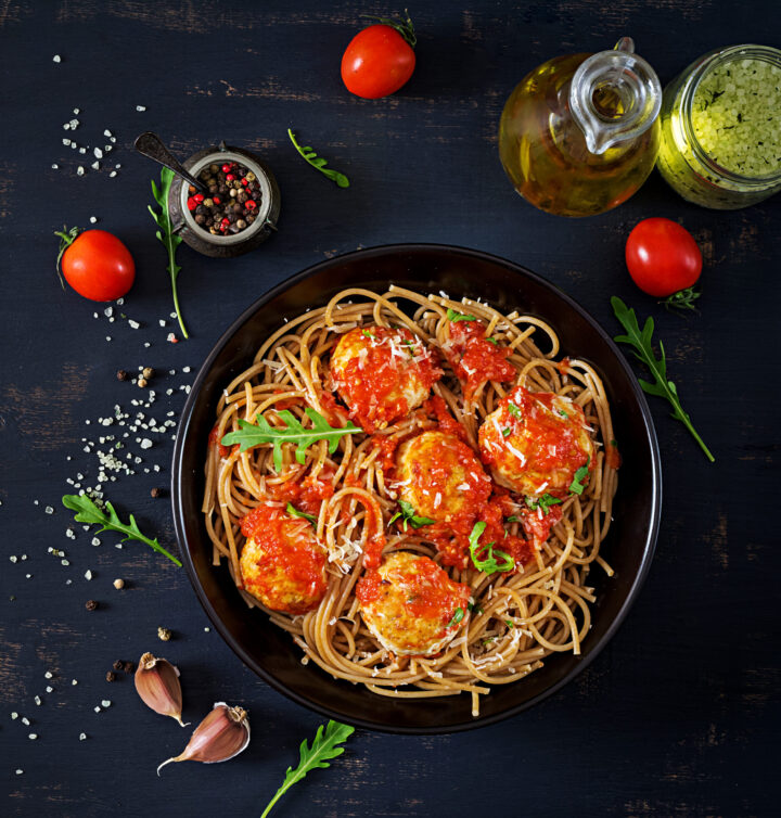 Eine schwarze Schale, gefüllt mit Spaghetti und Fleischbällchen mit Tomatensauce und Rucola, umgeben von Kirschtomaten, Knoblauchzehen, Olivenöl, Pfeffer und Kräutern auf einer dunklen Tischplatte - köstlich inspiriert von den besten Zutaten von WASGAU.
