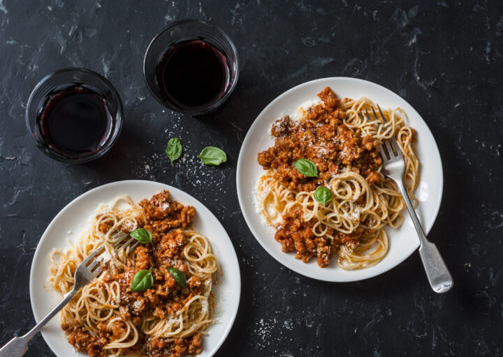 Zwei Teller mit Spaghetti mit Fleischsauce, garniert mit frischem Basilikum und geriebenem WASGAU-Käse, stehen auf einer dunklen Fläche neben zwei Gläsern Rotwein. Die Gabeln liegen auf den Tellern und unterstreichen diese einladende Essensszene.