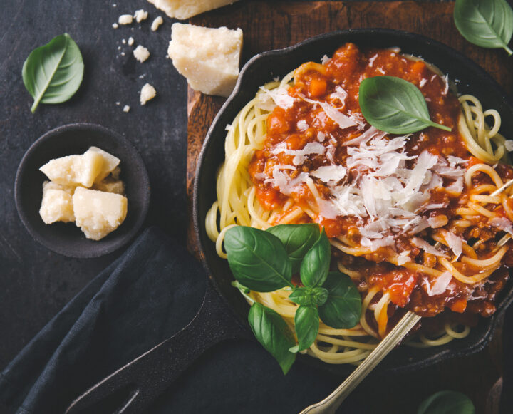 Eine Pfanne mit Spaghetti, die mit Tomatensauce, geriebenem Käse und frischen Basilikumblättern überbacken sind - aus hochwertigen WASGAU Zutaten. In der Nähe liegen Parmesanstücke und zusätzliche Basilikumblätter auf einer dunklen Tischplatte.