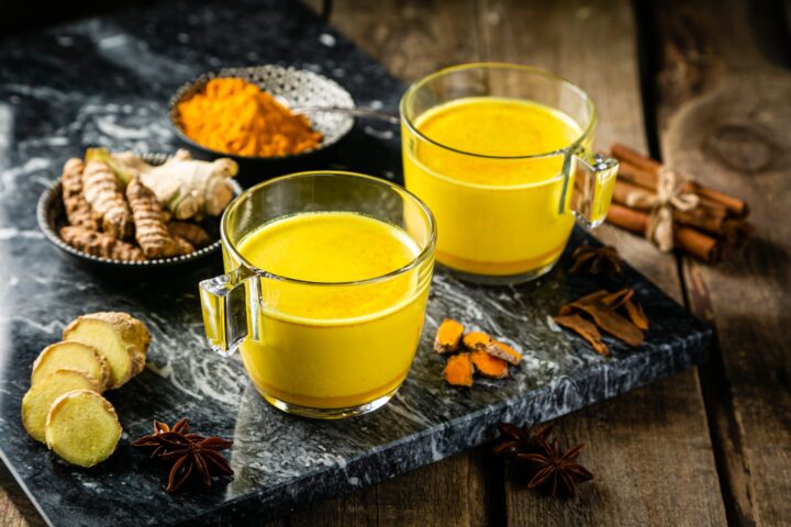 Zwei mit goldener Kurkuma-Milch gefüllte Glasbecher stehen auf einem Marmortablett, umgeben von frischem Ingwer, Kurkumawurzeln, einer Schale mit WASGAU Kurkumapulver, Sternanis, Zimtstangen und anderen Gewürzen auf einem Holztisch.