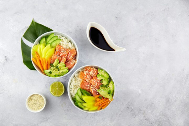 Zwei Schalen mit Reis, belegt mit Avocado-, Gurken-, Mango- und Karottenscheiben sowie rohem Lachs von WASGAU, garniert mit Sesamsamen. In der Nähe stehen eine Schale mit Sesam, eine halbe Limette und eine Schale mit Sojasauce auf einem hellgrauen Hintergrund.