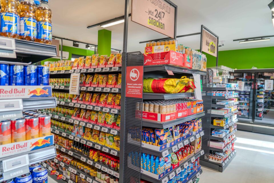 Ein Supermarktgang mit Snacks, Chips und Getränken in Regalen bei heller Beleuchtung.