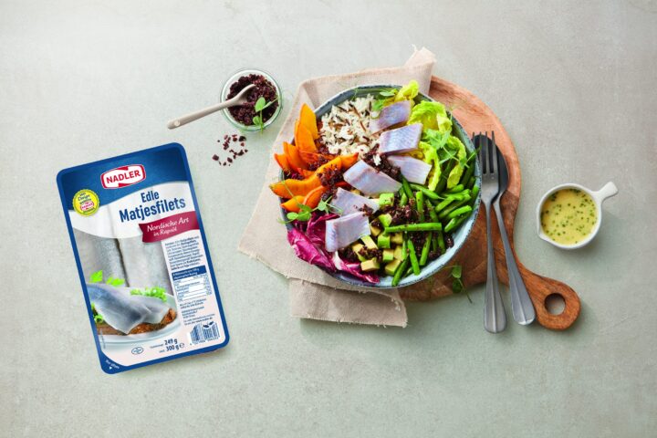 Eine Schale mit Heringsfilets, Reis, Salat, Karotten, Bohnen, Avocado und Rotkohl auf einem Holzbrett, daneben eine kleine Schale mit Dressing und eine Packung WASGAU Nadler Heringsfilets.