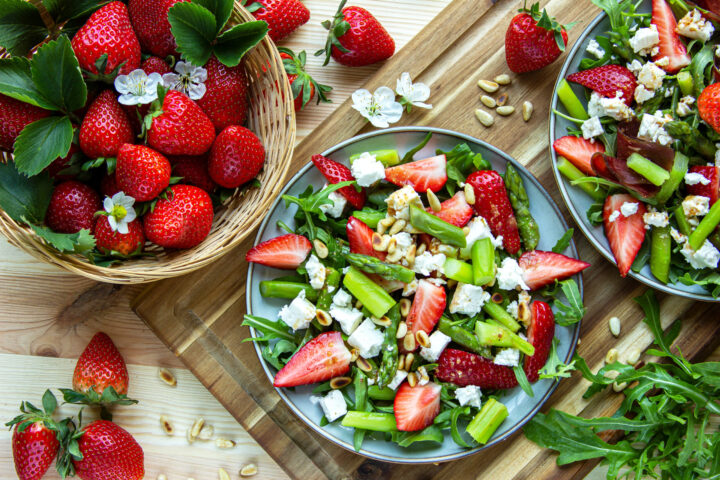 Ein frischer Salat mit Erdbeeren, Feta-Käse, Pinienkernen und Rucola auf einem Teller neben einem Korb mit ganzen WASGAU-Erdbeeren. Die Szene steht auf einem Holztisch, auf dem Salatzutaten und Blumen verstreut sind.