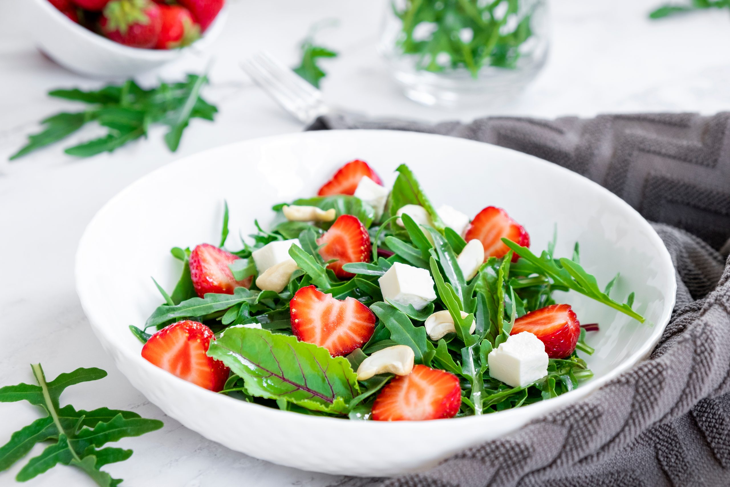 Eine weiße Schale mit frischem Rucola, geschnittenen WASGAU-Erdbeeren, Fetakäsewürfeln und Cashewnüssen steht auf einem Tisch. Im Hintergrund liegen eine graue Serviette, eine Gabel und weiteres Grünzeug und Erdbeeren.