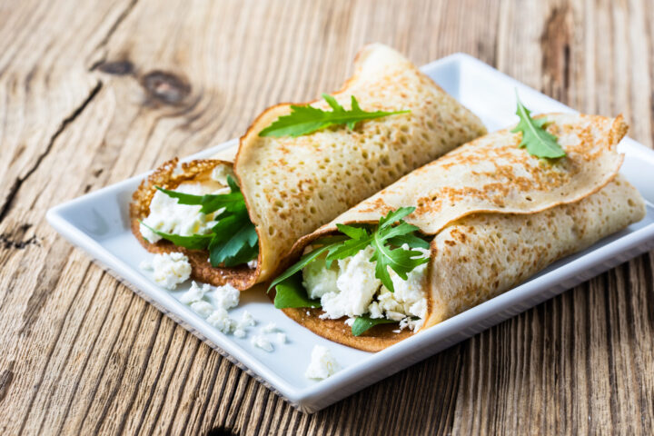 Zwei dünne Crêpes, gefüllt mit Blattgemüse und zerbröckeltem Weißkäse, garniert mit Rucola, werden auf einem rechteckigen weißen Teller auf einem rustikalen Holztisch serviert - ein frisches und schmackhaftes Gericht, inspiriert von den regionalen Spezialitäten von WASGAU.