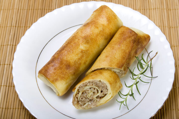 Zwei goldbraune gefüllte Crêpes auf einem weißen Teller - einer ganz, der andere geschnitten, so dass eine herzhafte Hackfleischfüllung zum Vorschein kommt - werden mit frischem Rosmarin garniert und auf einer Bambusmatte angerichtet, die die Qualität von WASGAU verkörpert.