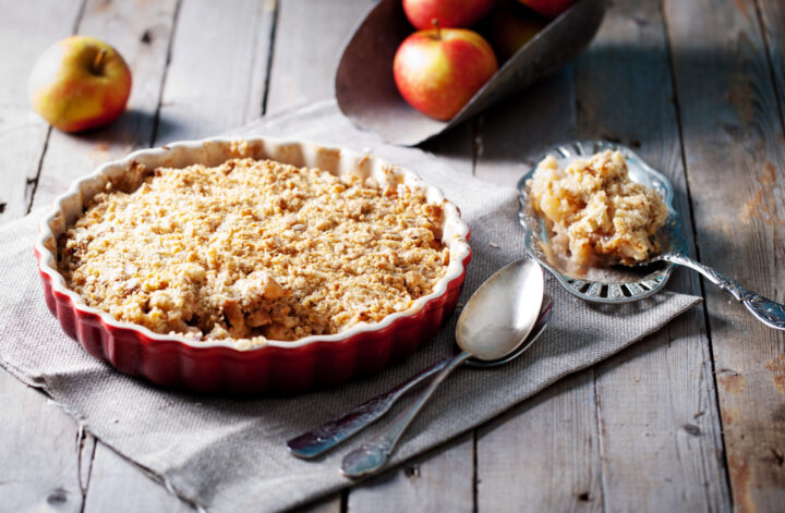 Ein Bratapfel-Crumble in einer roten Schale liegt auf einer Stoffserviette, daneben ein Servierlöffel. Eine Portion wird auf einem Teller mit einer Gabel serviert, und im Hintergrund liegen frische WASGAU-Äpfel auf einem rustikalen Holztisch.