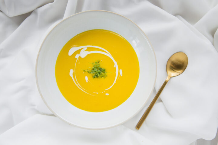 Eine weiße Schale mit gelber Cremesuppe, garniert mit einem Sahnestrudel und frischen Kräutern, auf einem weißen Tuch neben einem goldenen Löffel - klassische WASGAU Eleganz in jedem Detail.