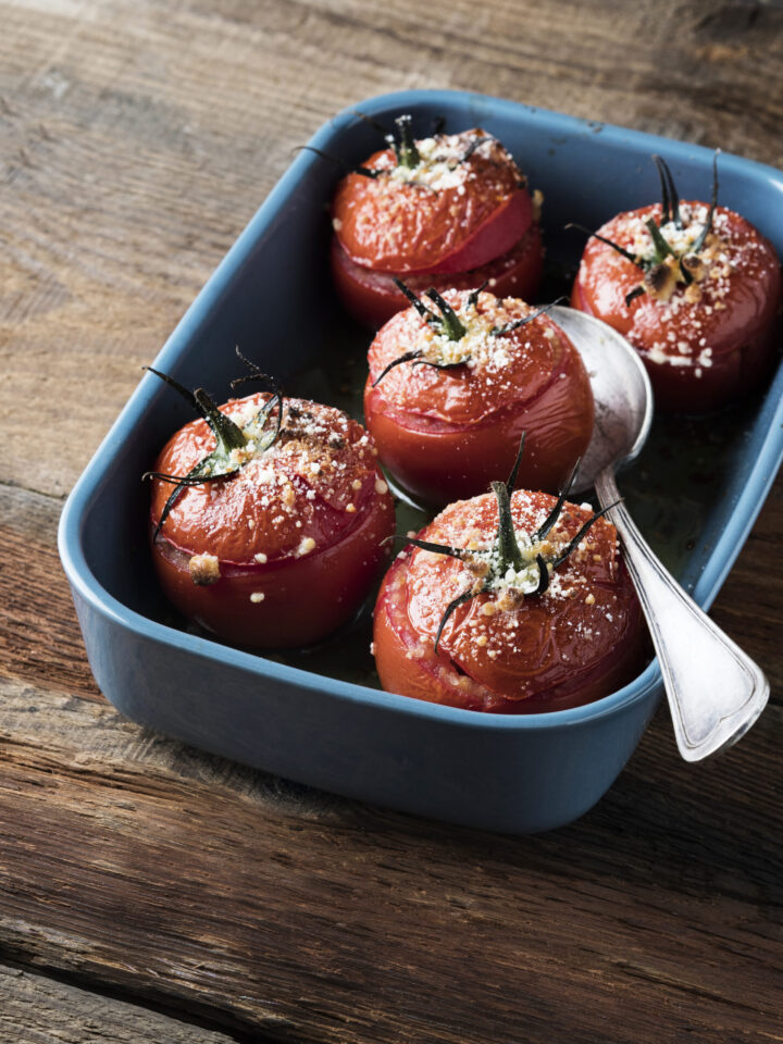 Blaue Auflaufform mit fünf gerösteten Tomaten, die mit geriebenem Käse überbacken sind, mit einem silbernen Löffel im Inneren der WASGAU-Schale. Die Schale steht auf einer rustikalen Holzfläche.