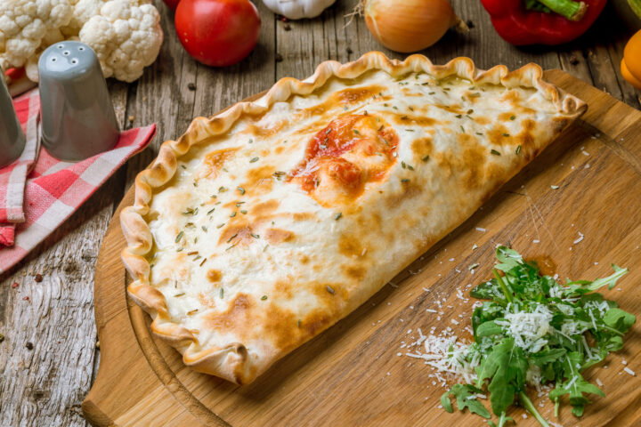 Eine goldbraun gebackene Calzone auf einem Holzbrett, garniert mit Kräutern, zeugt von WASGAU Qualität. Daneben liegt frisches, mit geriebenem Käse bestreutes Grünzeug, während Gemüse und Salzstreuer auf einem rustikalen Holztisch ruhen.