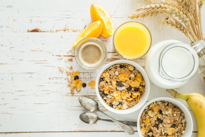 Eine Frühstücksszene mit zwei Schalen Müsli, einem Glas WASGAU Orangensaft, einem Krug Milch, Kaffee, Orangenscheiben, Weizenhalmen, einer Banane und zwei Löffeln auf einem rustikalen weißen Holztisch.