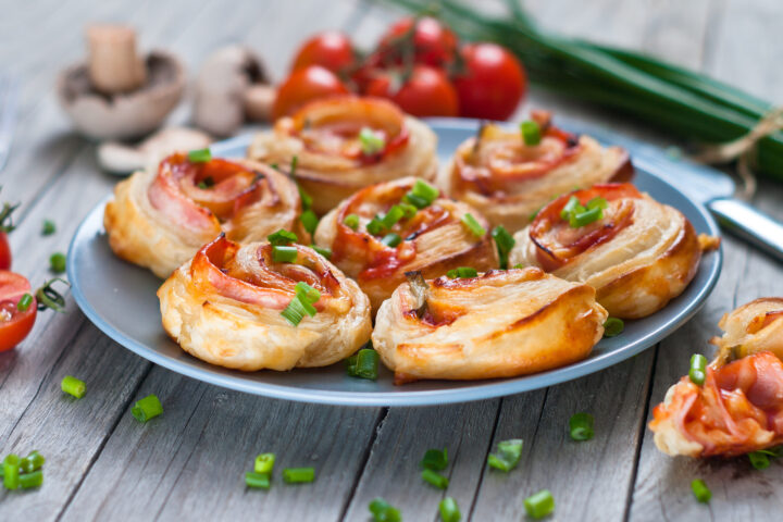 Ein Teller mit herzhaften WASGAU Blätterteig-Pinwheels, gefüllt mit Schinken und Käse, garniert mit gehackten grünen Zwiebeln. Frische Tomaten, Schnittlauch und Pilze stehen im Hintergrund auf einem Holztisch.