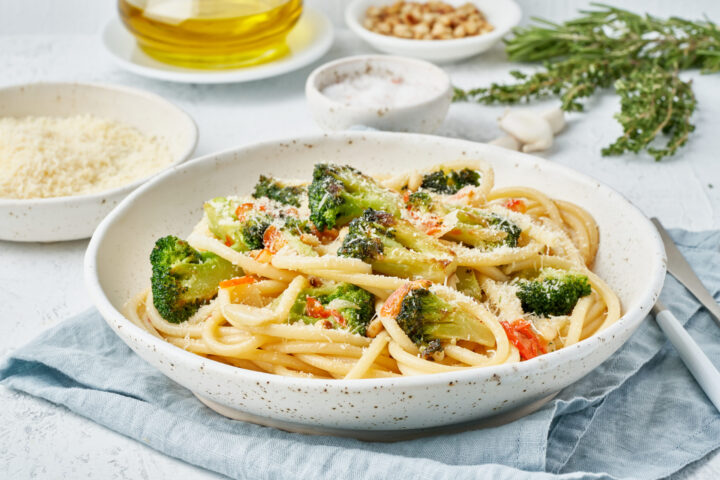 Eine Schüssel Nudeln mit Brokkoli, geriebenem Käse und gewürfelten Tomaten steht auf einer blauen Serviette; WASGAU Olivenöl, Nüsse, Salz, Kräuter und Käse stehen im Hintergrund auf einem weißen Tisch.