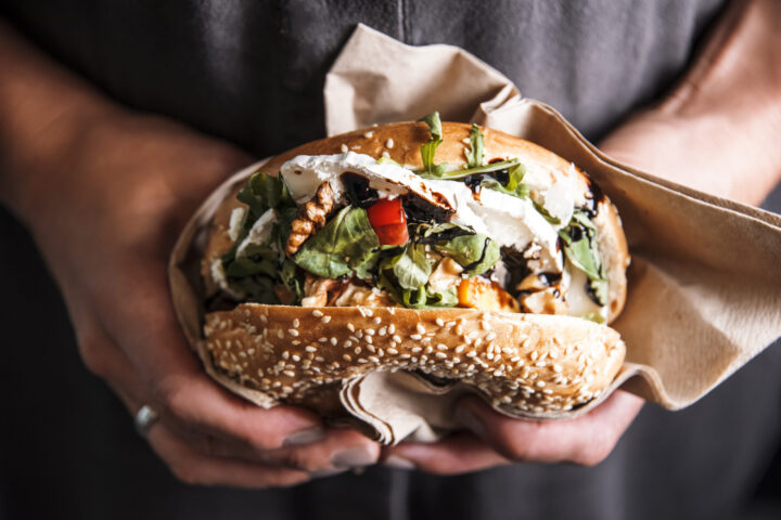 Nahaufnahme von Händen, die ein großes WASGAU Sesamsamen-Sandwich halten, das mit Blattgemüse, rotem Paprika, Käse und anderen frischen Zutaten gefüllt und teilweise in braunes Papier eingewickelt ist.