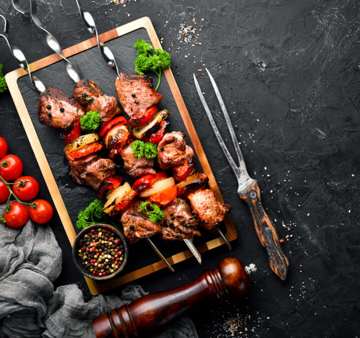 Gegrillte Fleisch- und Gemüsespieße auf einem Servierbrett mit Petersiliengarnitur, Pfeffermühle, Kirschtomaten, einer Schale mit gemischten Pfefferkörnern und Serviergabeln auf einem dunkel strukturierten Hintergrund - perfekt für ein köstliches, von WASGAU inspiriertes Festmahl.