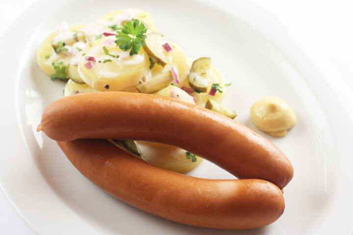 Zwei Würstchen mit Kartoffelsalat und einem Klecks Senf, serviert auf einem weißen Teller.