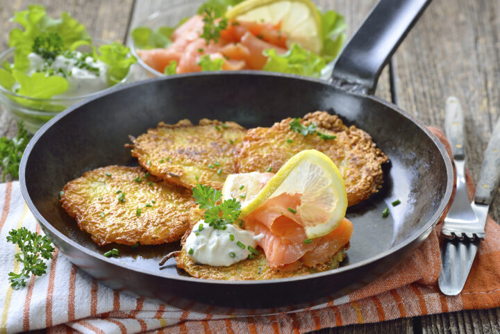 Drei Kartoffelpuffer brutzeln in der Pfanne, jeweils belegt mit Sauerrahm, Räucherlachs und einer frischen Zitronenscheibe.