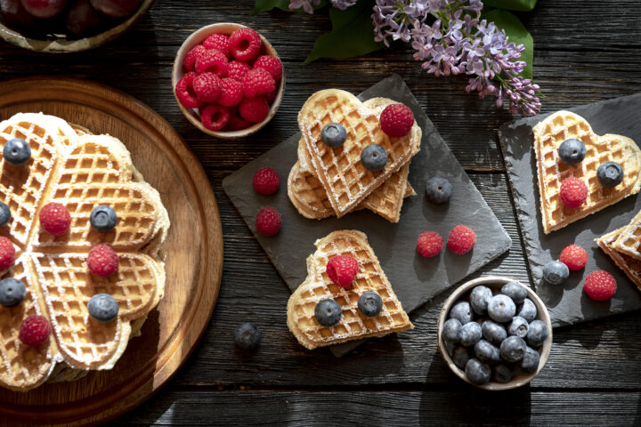 Herzförmige Waffeln, belegt mit Himbeeren und Heidelbeeren, serviert mit frischen Beeren auf einem rustikalen Tisch.