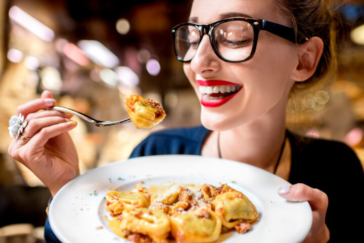 Lächelnde Frau mit rotem Lippenstift und Brille, die einen Teller mit WASGAU Tortellini-Nudeln in der Hand hält, um einen Bissen zu essen, in einem warm beleuchteten Innenraum.