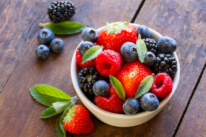 Auf einem rustikalen Holztisch steht eine weiße Schale mit frischen WASGAU-Erdbeeren, Heidelbeeren, Brombeeren und Himbeeren, garniert mit Minzblättern. Ein paar Beeren und Minzblätter sind um die Schale herum verstreut.