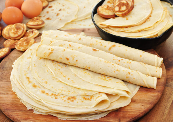 Ein Stapel dünner, goldbrauner WASGAU Crêpes liegt auf einem Holzbrett, darauf einige gerollte Crêpes. Im Hintergrund befinden sich Eier, Mini-Pfannkuchen und weitere Crepes in einer schwarzen Pfanne.