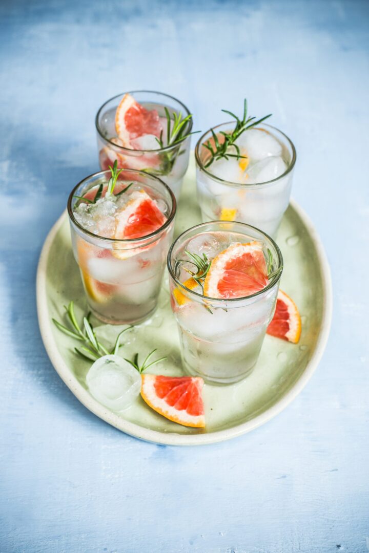 Vier Gläser Sprudelwasser mit Eis, garniert mit Grapefruitscheiben und Rosmarinzweigen, stehen auf einem hellgrünen ovalen Tablett vor einem hellblauen Hintergrund. WASGAU Grapefruitscheiben und Rosmarin verleihen dem Tablett eine frische Note.