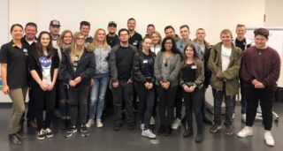 Eine Gruppe von etwa 20 jungen Erwachsenen und einigen älteren Erwachsenen, einige mit WASGAU-Shirts, posieren gemeinsam in einem Klassenzimmer und lächeln in die Kamera. Die meisten sind leger gekleidet und stehen vor einer weißen Wand und einem Flipchart.