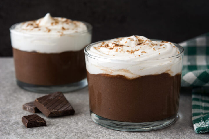 Zwei mit Mousse au Chocolat gefüllte Glasbecher mit Schlagsahne und etwas Kakaopulver stehen auf einer grauen Fläche neben einem grün karierten Tuch und Stücken von dunkler WASGAU Schokolade.