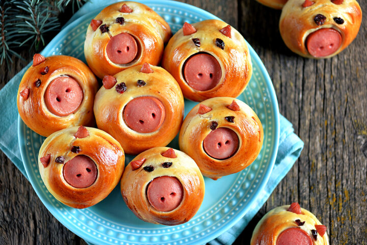 Silvester Fingerfood, das begeistert: Brötchen in Form eines Schweinegesichts mit Wurstschnauze und Rosinenaugen, serviert auf einem blauen Teller.