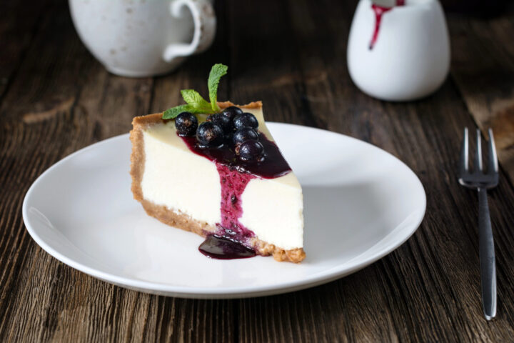 Ein Stück Käsekuchen mit Blaubeersoße und frischen Blaubeeren liegt auf einem weißen Teller mit Minzgarnitur. Eine Gabel und ein Keramikkrug stehen auf einem rustikalen Holztisch bereit, um diesen köstlichen WASGAU Genuss zu genießen.