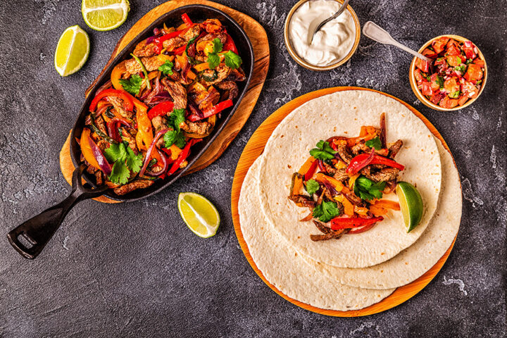 Fajita-Mischung mit Paprika und Rindfleisch in einer Pfanne, Tortillas, Limettenspalten, Pico di Gallo, Salsa und saure Sahne auf einer dunklen Fläche.
