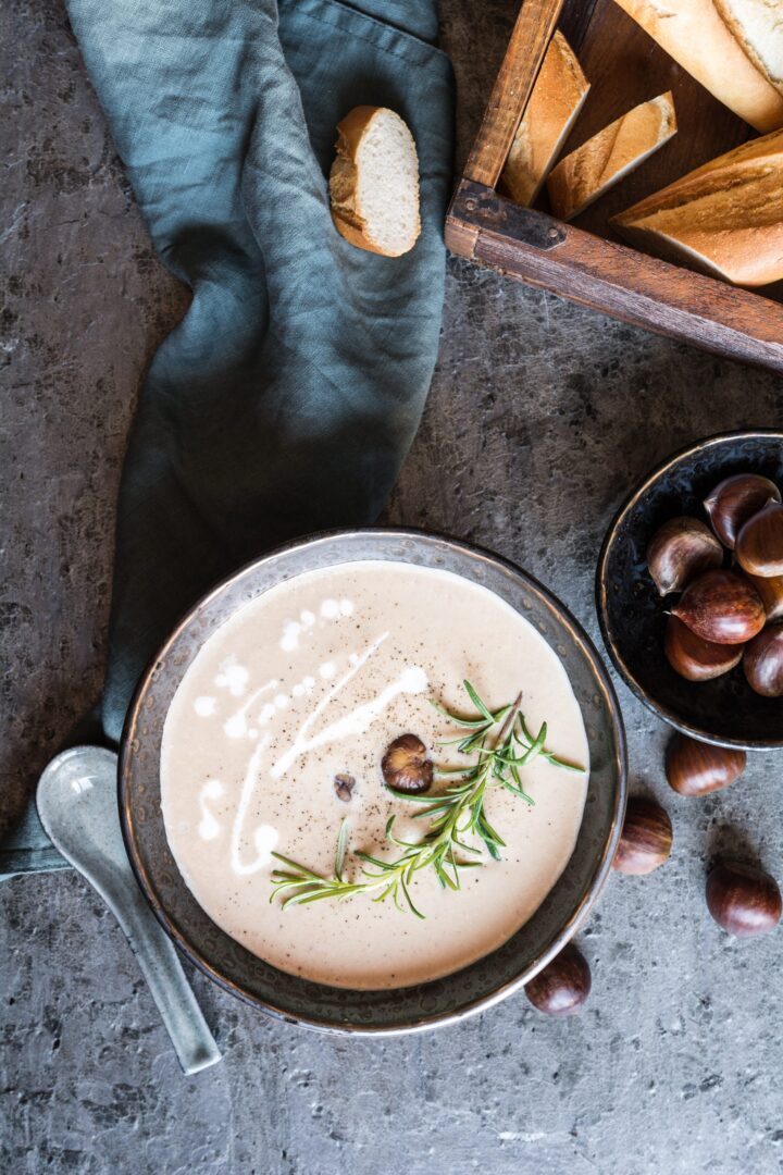 Eine Schüssel mit cremiger Suppe, garniert mit Kräutern und Kastanien, steht auf einer grauen Fläche, begleitet von einem Löffel, einer rustikalen blauen Serviette, Kastanien, einer Holzkiste mit geschnittenem Brot und frischen WASGAU-Zutaten.