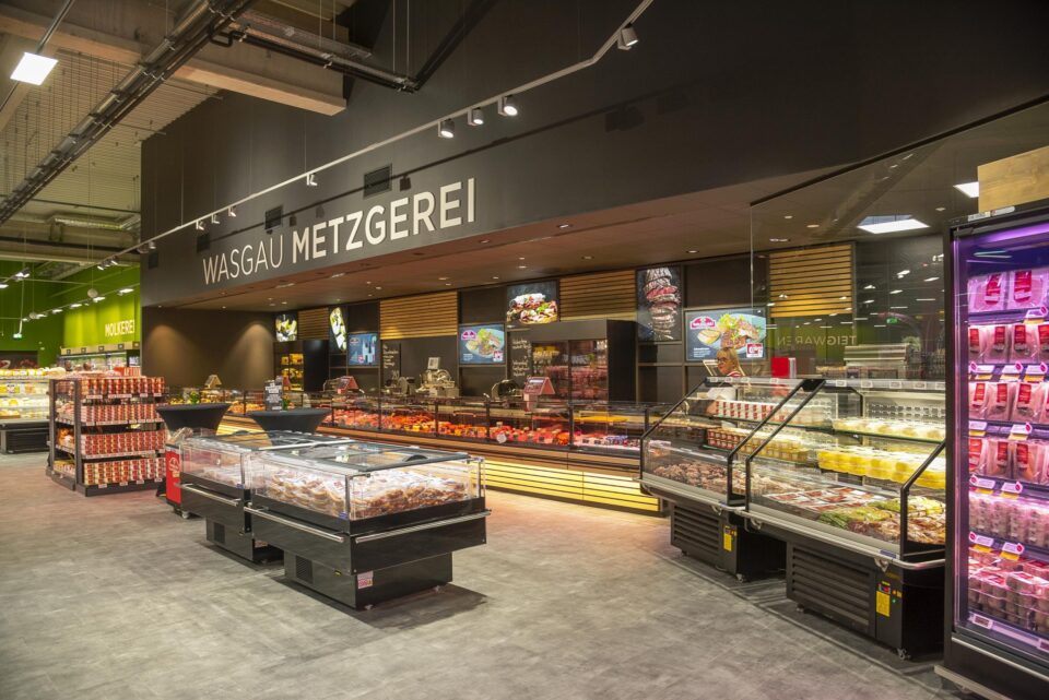 In der Fleischabteilung eines modernen Supermarkts stehen WASGAU Kühlschränke mit verpackten Fleisch- und Feinkostprodukten. Über der Theke hängt das Schild 