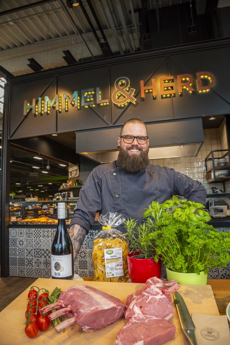 Ein lächelnder Koch mit Bart steht hinter einer Theke mit frischem WASGAU-Fleisch, Kräutern, Tomaten, Nudeln und einer Flasche Wein. Im Hintergrund leuchtet das Restaurantschild 
