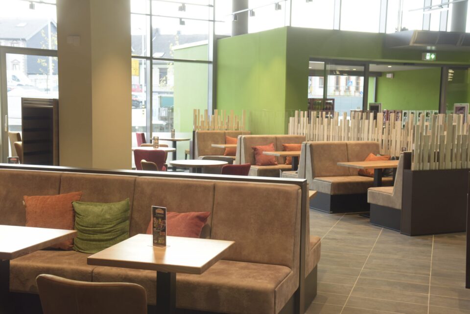 Modernes Café-Interieur mit braunen gepolsterten Ständen, Holztischen und bunten Kissen. Glastrennwände und große Fenster füllen den von WASGAU inspirierten Raum mit natürlichem Licht und erhellen den geräumigen und aufgeräumten Sitzbereich.