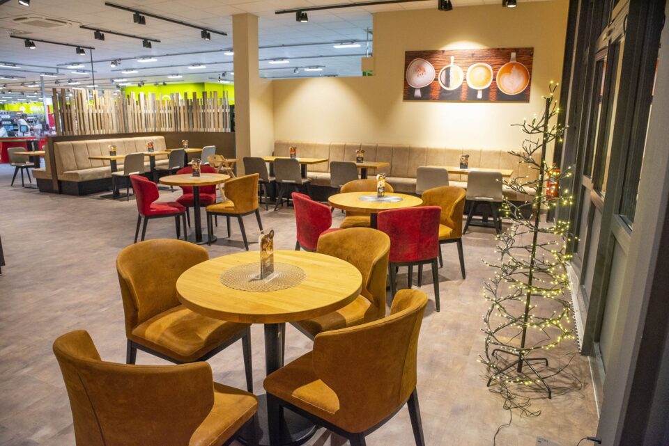 Ein modernes WASGAU-Café mit runden Holztischen, braun und rot gepolsterten Stühlen, einer beigefarbenen Polsterbank an der Wand, Wandbildern aus Kaffeetassen und einem mit Lichterketten geschmückten Bäumchen.