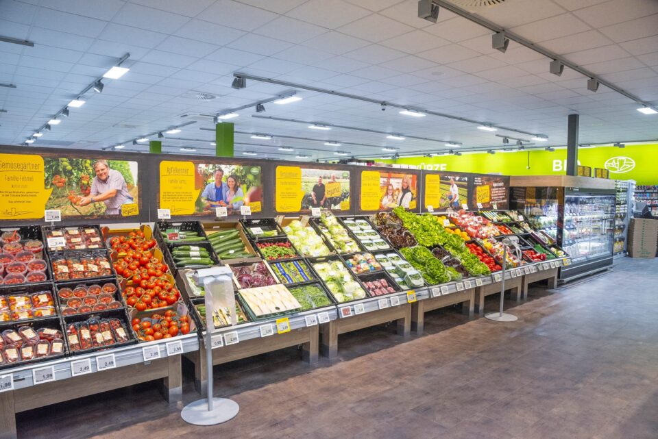 In der Obst- und Gemüseabteilung des WASGAU-Supermarkts sind verschiedene frische Obst- und Gemüsesorten, darunter Tomaten, Gurken, Salat und Kräuter, übersichtlich in Behältern unter heller Beleuchtung und mit Informationsschildern darüber angeordnet.