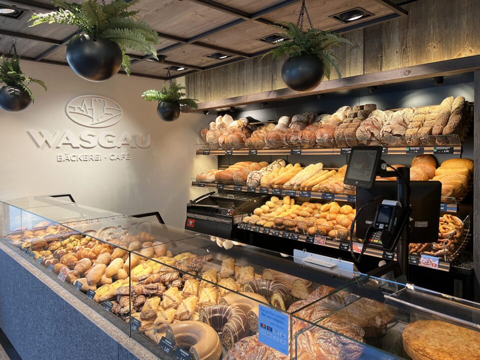 Eine WASGAU Bäckerei mit Regalen voller Brotsorten und Gebäck hinter einer Glastheke. Das moderne Geschäft ist mit hängenden Pflanzen und einem "WASGAU Bäckerei - Café" Logo an der Wand ausgestattet.