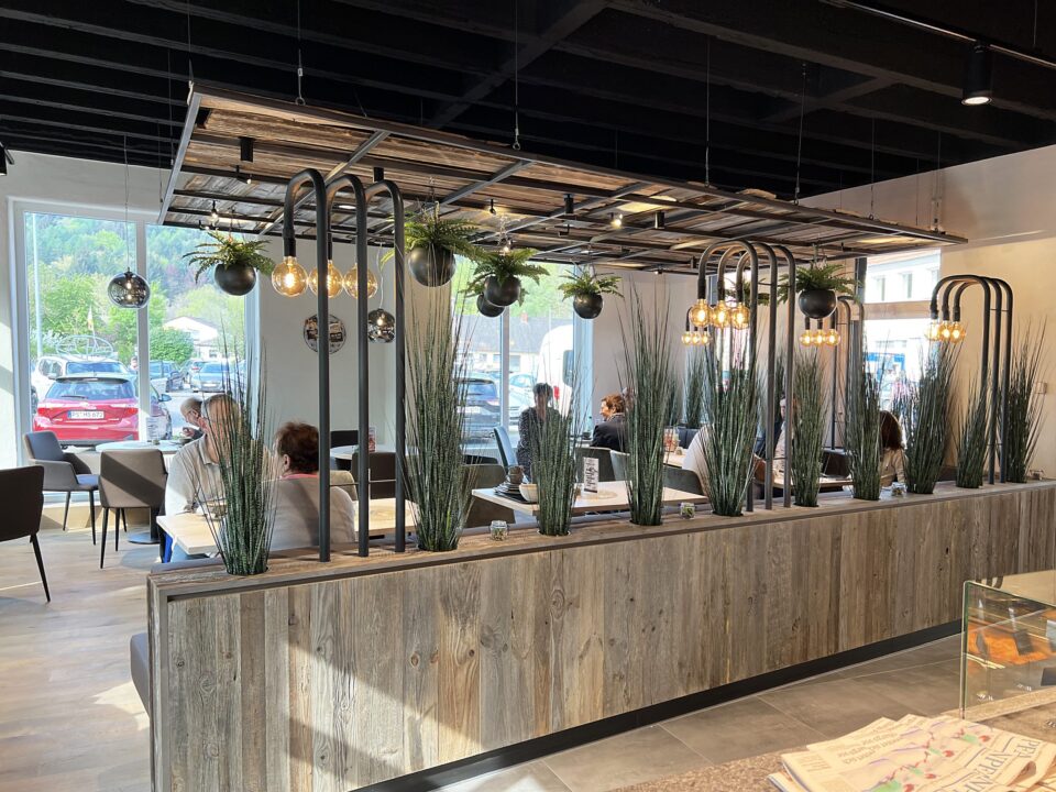 Modernes Café-Interieur mit Holzdekor, hängenden Pflanzen und Kugel-Pendelleuchten. Die Gäste sitzen an Tischen vor großen Fenstern, von denen aus man Autos und Bäume sehen kann. WASGAU Pflanzgefäße mit hohem Gras unterteilen den Sitzbereich.