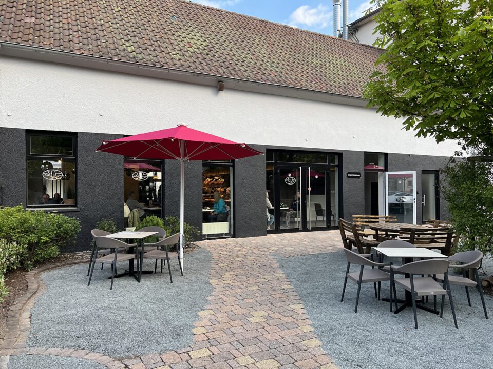 Außensitzbereich des WASGAU Cafés mit grauen Tischen und Stühlen, einem roten Sonnenschirm und einem gemauerten Gehweg. Das Café hat große Fenster, eine Glastür und ein weißes und dunkelgraues Äußeres. Der einladende Innenhof ist von Grün umgeben.