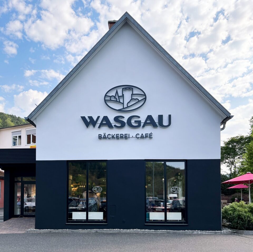 Ein modernes weiß-schwarzes WASGAU Gebäude mit großen Fenstern beherbergt das "WASGAU Bäckerei-Café". Das Logo über dem Eingang zeigt ein Schloss und Hügel. Der Himmel ist teilweise bewölkt, und es gibt Sitzgelegenheiten im Freien mit einem rosa Regenschirm.