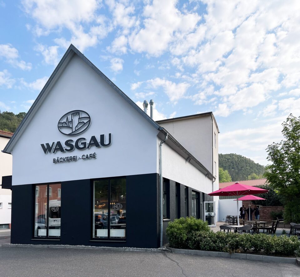 Ein modernes weißes Gebäude mit schwarzen Akzenten beherbergt ein WASGAU Bäckerei-Café. Große Fenster zeigen zur Straße, und ein einladender Außenbereich mit Tischen und rosafarbenen Sonnenschirmen empfängt die Gäste unter einem teilweise bewölkten Himmel.