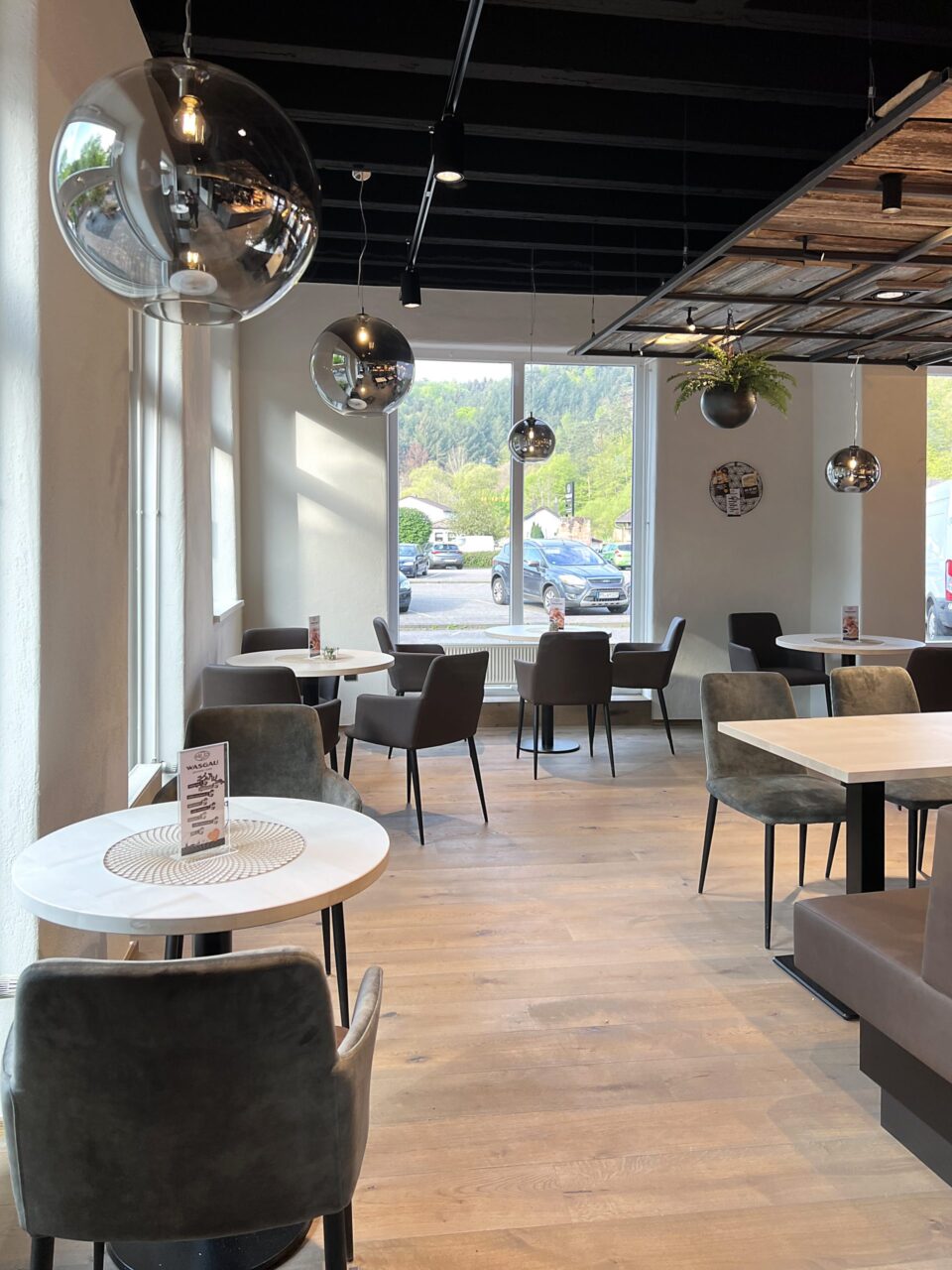 Ein modernes, luftiges WASGAU-Café mit runden Tischen, grauen Stühlen, großen Fenstern und Kugel-Pendelleuchten. Natürliches Licht erfüllt den Raum, mit Blick auf grüne Bäume und geparkte Autos draußen.