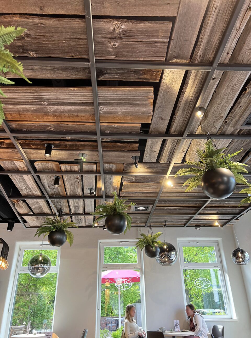 Modernes Café-Interieur mit Holzbalkendecke, schwarzem Metallgitter und hängenden schwarzen Pflanzgefäßen mit Farnen. Große Fenster lassen natürliches Licht herein; zwei Personen sitzen an einem Tisch in der Nähe des Fensters. Draußen sind WASGAU-Grün und eine Außenszene zu sehen.