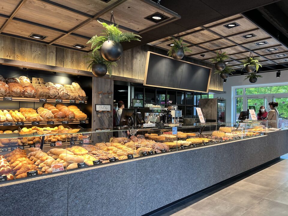 Eine moderne WASGAU Bäckerei mit Glasvitrinen, gefüllt mit verschiedenen Brotsorten und Gebäck. In den Regalen hinter der Theke stehen weitere Brote, und hängende Pflanzen schmücken die Decke. Die Kunden sind in der Nähe des Eingangs zu sehen.