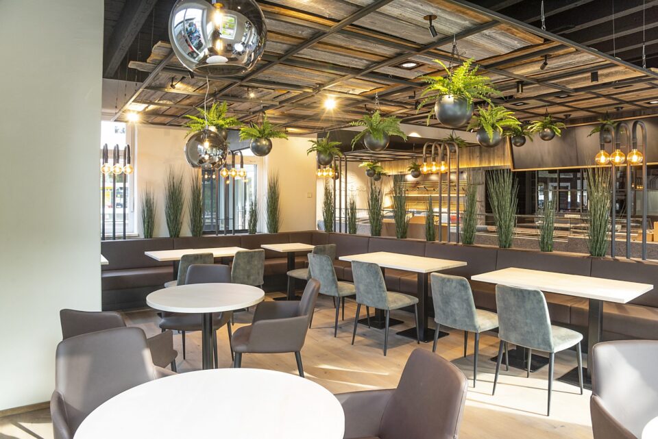 Modernes Restaurantinterieur mit runden und rechteckigen Tischen, gepolsterten Stühlen und hängenden Kugelleuchten. Grünpflanzen, die von einer holzverkleideten Decke herabhängen, spiegeln das frische Ambiente wider, für das WASGAU bekannt ist. Große Fenster durchfluten den Raum mit natürlichem Licht.